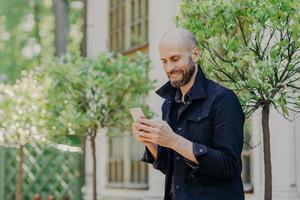 positivo jovem empresário careca com barba escura espessa, feliz em receber mensagem de felicitações, comemora aniversário, posa contra a bela vista do parque. conceito de pessoas e tecnologias modernas foto