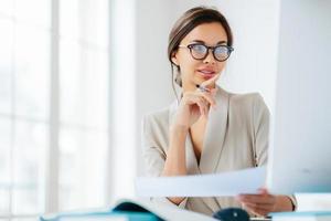 economista feminina posa no espaço de coworking, focada no monitor, verifica ou verifica os números do relatório financeiro, segura documento em papel e caneta, tem cabelos escuros penteados, usa terno formal foto