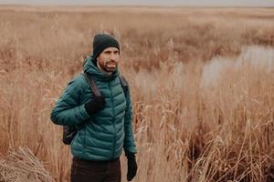 tiro ao ar livre de viajante masculino pensativo vestido com roupas quentes, de bom humor, tem mochila nas costas, caminha em ambiente rural perto de campo durante o outono, goza de uma atmosfera calma. conceito de lazer foto