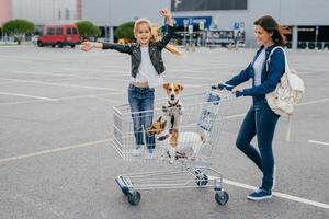 conceito de compras e família. feliz mulher carrega carrinho em que garotinha fica com cachorros, posar contra o fundo do shopping, alegrar novas compras. pessoas, felicidade, conceito de dia de folga foto