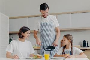 ocupado pai carinhoso prepara delicioso café da manhã para esposa e filha, usa avental, coloca ovos fritos no prato. alegre mãe e filho conversam enquanto se sentam à mesa da cozinha, fazem a refeição foto