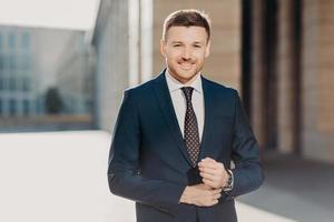 homem bonito com barba, vestido de terno formal, posa ao ar livre, tem relógio no braço, estando em alto astral após contrato assinado com sucesso, sente-se confiante e satisfeito. sorrisos de empresário próspero foto