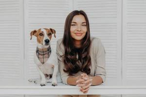 foto de mulher bonita com cabelos escuros, passa o tempo de lazer com o cachorro jack russel terrier, adora animais, tem um relacionamento amigável com o animal de estimação, posam juntos na mesa branca, brincam juntos.