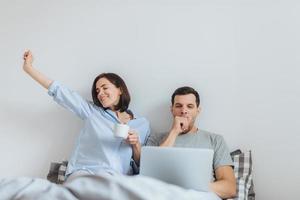 mulher feliz se estende na cama, bebe café quente e seu marido trabalha a noite toda no computador portátil, boceja como quer dormir, passa tempo juntos no quarto, isolado sobre fundo branco foto