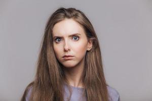 tiro na cabeça de mulher bonita com olhos azuis e pele pura, olha séria e com raiva para a câmera, tem cabelo liso escuro, isolado sobre fundo cinza. tiro horizontal de linda jovem interior foto