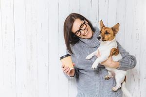 modelo feminino morena feliz usa suéter de malha quente, óculos, carrega seu cachorro favorito e café para viagem, vai passear juntos, ter bons relacionamentos amigáveis. conceito de pessoas e animais de estimação foto