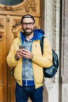 jovem viajante em roupas da moda em pé no museu tendo um olhar feliz por estar lá. macho barbudo alegre na jaqueta amarela segurando a mochila nas costas segurando o celular visitando o museu regional foto