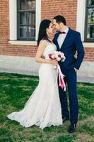 afetuoso casamento jovem casal vai se beijar, sendo feliz em celebrar seu casamento. linda mulher morena de vestido branco e noivo bonito terno preto elegante. conceito de união foto