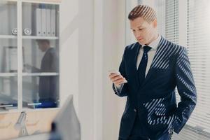 diretor masculino atraente vestido com roupas corporativas, verifica e-mail no celular, fica perto da janela com obturador em seu armário aconchegante, blogs site, compartilha notícias com colegas, usa internet foto