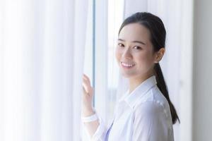 mulher bonita asiática tem cabelo comprido preto na camisa branca. ela está sorrindo e de pé perto da janela com cortina branca. foto