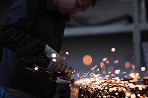 interior da fábrica de engenharia da indústria pesada com trabalhador industrial usando rebarbadora e cortando um tubo de metal. empreiteiro em uniformes de segurança e fabricação de capacetes de estruturas metálicas. foto