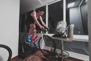 homem andando de bicicleta no treinador de máquina ele está se exercitando em casa à noite jogando jogo de corrida de bicicleta online foto