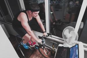 homem andando de bicicleta no treinador de máquina ele está se exercitando em casa à noite jogando jogo de corrida de bicicleta online foto