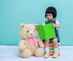 criança menina asiática ou uma menina asiática bonita lendo um livro no fundo verde e sentado nos livros com um ursinho de pelúcia de brinquedo foto