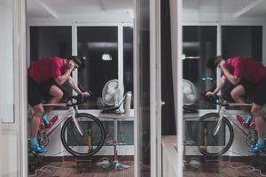 homem andando de bicicleta no treinador de máquina ele está se exercitando em casa à noite jogando jogo de corrida de bicicleta online foto