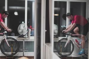 homem andando de bicicleta no treinador de máquina ele está se exercitando em casa à noite jogando jogo de corrida de bicicleta online foto