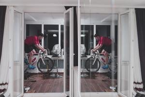 homem andando de bicicleta no treinador de máquina ele está se exercitando em casa à noite jogando jogo de corrida de bicicleta online foto