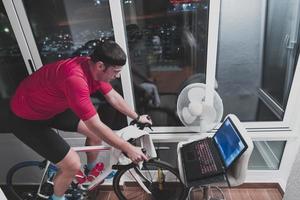 homem andando de bicicleta no treinador de máquina ele está se exercitando em casa à noite jogando jogo de corrida de bicicleta online foto