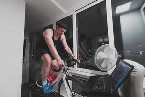 homem andando de bicicleta no treinador de máquina ele está se exercitando em casa à noite jogando jogo de corrida de bicicleta online foto
