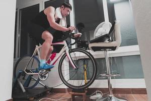 homem andando de bicicleta no treinador de máquina ele está se exercitando em casa à noite jogando jogo de corrida de bicicleta online foto