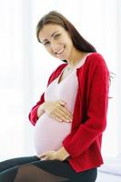 mulher grávida senta-se na cama em seu quarto, acariciando sua barriga com um rosto feliz e sorridente. foto