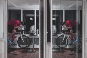 homem andando de bicicleta no treinador de máquina ele está se exercitando em casa à noite jogando jogo de corrida de bicicleta online foto