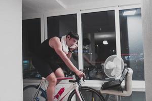 homem andando de bicicleta no treinador de máquina ele está se exercitando em casa à noite jogando jogo de corrida de bicicleta online foto