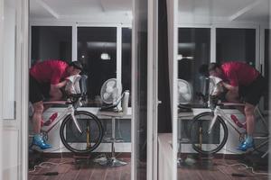 homem andando de bicicleta no treinador de máquina ele está se exercitando em casa à noite jogando jogo de corrida de bicicleta online foto