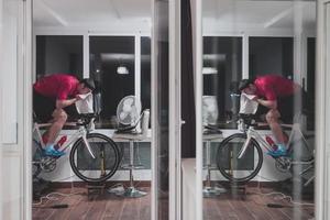 homem andando de bicicleta no treinador de máquina ele está se exercitando em casa à noite jogando jogo de corrida de bicicleta online foto