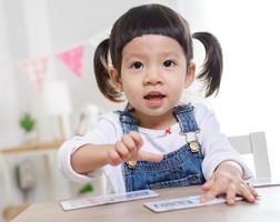 menina asiática sentada à mesa na sala, menina pré-escolar jogando o quebra-cabeça em dia ensolarado, jardim de infância ou creche. foto