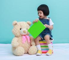 criança menina asiática ou uma menina asiática bonita lendo um livro no fundo verde e sentado nos livros com um ursinho de pelúcia de brinquedo