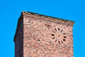 velha torre de tijolos vermelhos abandonada com relógio redondo na fachada, alvenaria vintage, fundo de céu azul foto