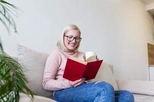 jovem em casa sentada no sofá moderno relaxando em sua sala lendo livro e bebendo café ou chá. cama aconchegante branca e uma linda garota, lendo um livro, conceitos de lar e conforto foto