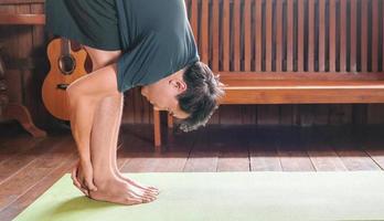 homem asiático desportivo de preto está fazendo ioga enquanto se exercita no tapete de ioga em casa com piso de madeira. foto