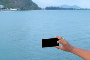 a tela preta do celular à mão com fundo desfocado do mar, o uso da imagem para selfie com grupo de amigos foto