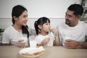 uma família tailandesa asiática saudável, uma filha pequena e pais jovens bebem leite branco fresco em vidro e pão alegria juntos em uma mesa de jantar de manhã, bem-estar nutrição casa café da manhã refeição estilo de vida. foto