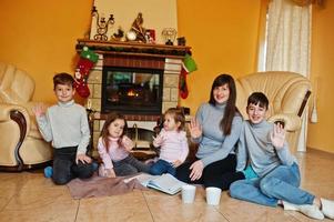 feliz jovem grande família por uma lareira na sala de estar quente em dia de inverno. mãe com quatro filhos em casa. foto