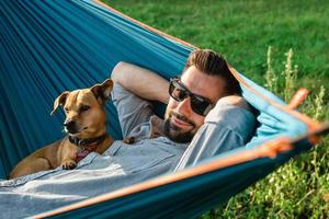 sorrindo atraente homem europeu em óculos de sol está descansando na rede com seu cachorrinho fofo. foto