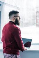 empresário pensativo em óculos, olhando pela janela enquanto trabalhava no laptop foto