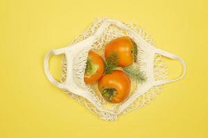 frutas de caqui estão no saco de malha branca em um fundo amarelo. conceito de comida saudável foto