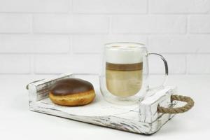 uma xícara de café com leite e uma rosquinha vitrificada em uma bandeja de madeira para o café da manhã cedo foto