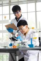 ciência, química, tecnologia, biologia e conceito de laboratório - cientista sênior asiático examina algo em uma placa de petri enquanto cientista júnior asiático tomando notas. foto