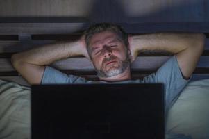 homem cansado e estressado atraente, viciado em trabalho, trabalhando tarde da noite, exausto na cama, ocupado com o computador portátil, sentindo-se sonolento e sobrecarregado em conceito de estresse de prazo de projeto de negócios