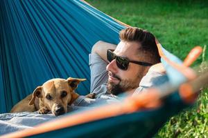 sorrindo atraente homem europeu em óculos de sol está descansando na rede com seu cachorrinho fofo. foto