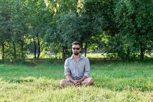 jovem europeu bonito em roupas casuais e óculos de sol, sentado em uma grama no parque de verão. foto