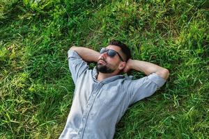 jovem homem europeu de esportes bonito em óculos de sol está descansando em uma grama no parque de verão, vista superior. foto