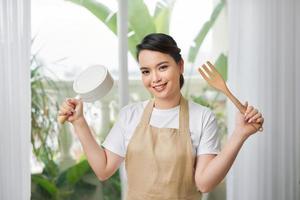 linda garota alegre cozinheira em roupas de cozinha segura uma frigideira e cozinha o jantar foto