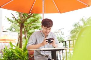 jovem macho sério sentado à mesa de madeira com câmera fotográfica e se preparando para sessão de fotos enquanto tomava café