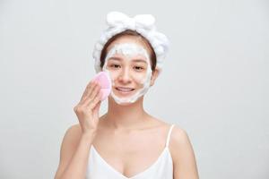 feliz jovem asiática aplicando espuma de limpeza no rosto e usando toalha na cabeça. foto