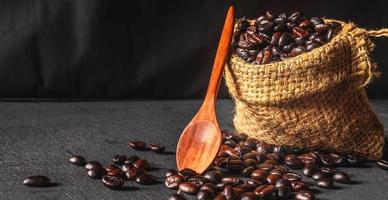 grãos de café torrados marrons do produto em um saco marrom com colher de pau em fundo preto de madeira foto